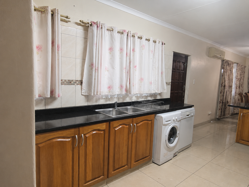 A functional kitchen featuring wooden cabinetry, granite countertops, a double sink, and integrated laundry and dishwasher appliances with neutral tiled flooring.