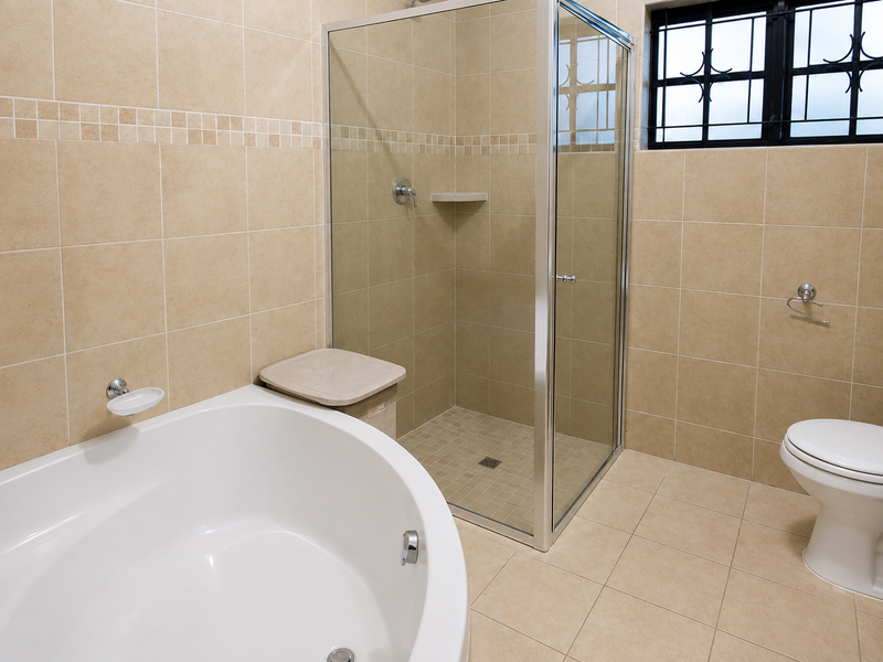 A clean, neutral-toned bathroom featuring a corner bathtub, a separate glass-enclosed shower, and a toilet, finished with floor-to-ceiling ceramic tiling.