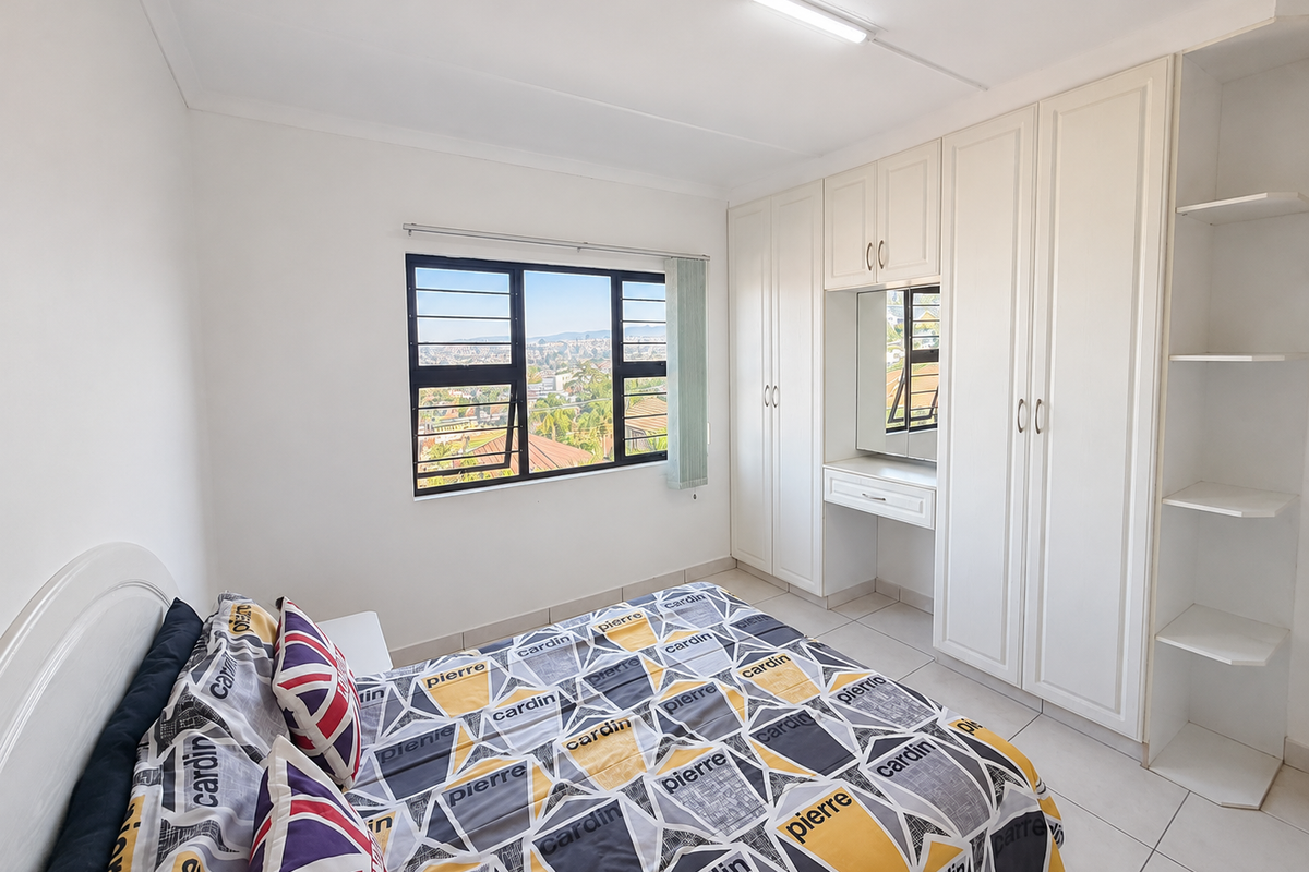 A bright bedroom featuring extensive white built-in cabinetry with an integrated vanity, tiled flooring, and large windows offering an elevated neighbourhood view.