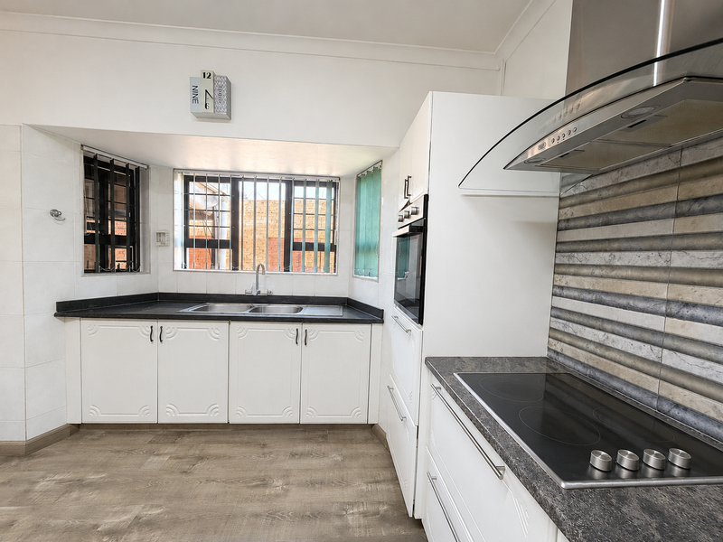 A clean, functional kitchen featuring white cabinetry, dark laminate countertops, a built-in oven, electric cooktop, and a double sink with security bars on windows.