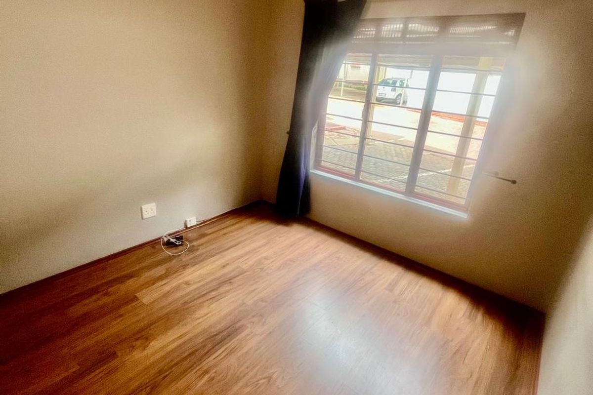 A bright, empty bedroom featuring warm-toned laminate flooring, neutral walls, and a window fitted with security bars.
