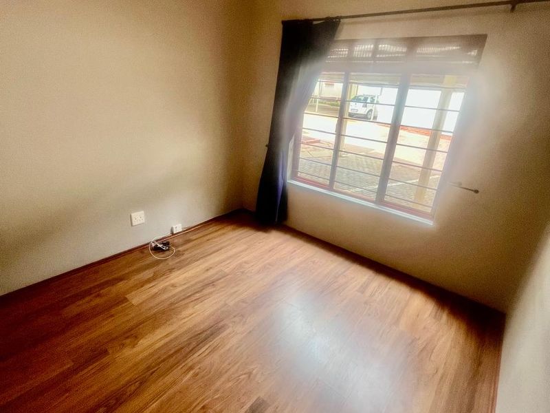 A bright, empty bedroom featuring warm-toned laminate flooring, neutral walls, and a window fitted with security bars.