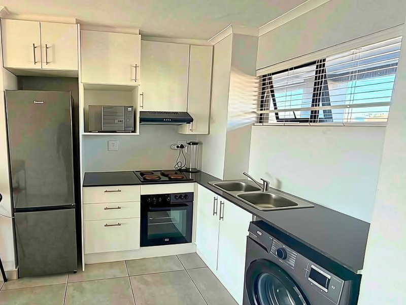 A compact, functional kitchen featuring white cabinetry, dark countertops, integrated oven and hob, and tiled flooring with natural light from the window.