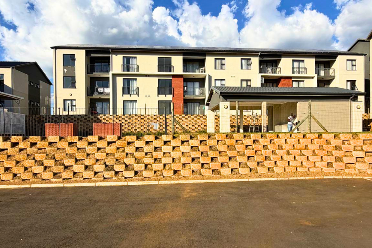Exterior view of a multi-story apartment complex featuring balconies, a retaining wall, and a paved parking area.