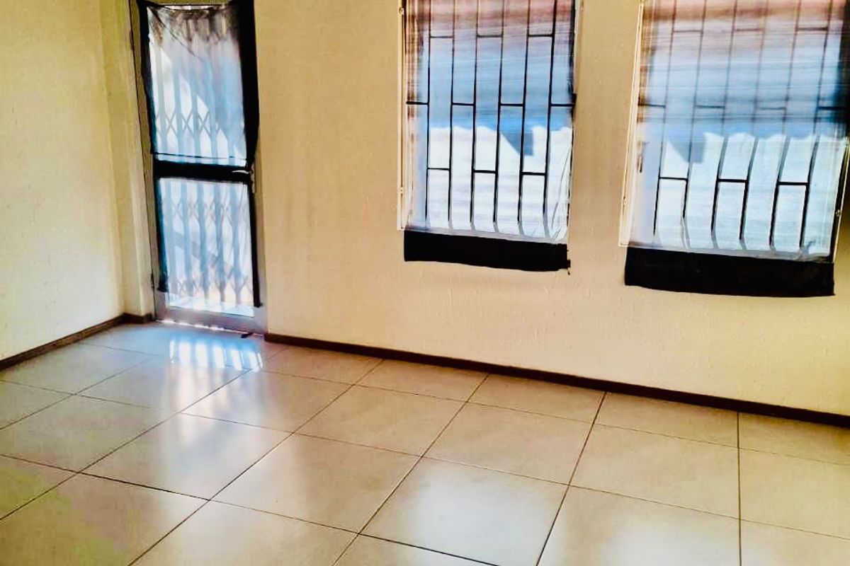 An empty living room featuring ceramic floor tiles, security gate, and burglar bars on the windows, requiring cosmetic updates to improve its overall condition.