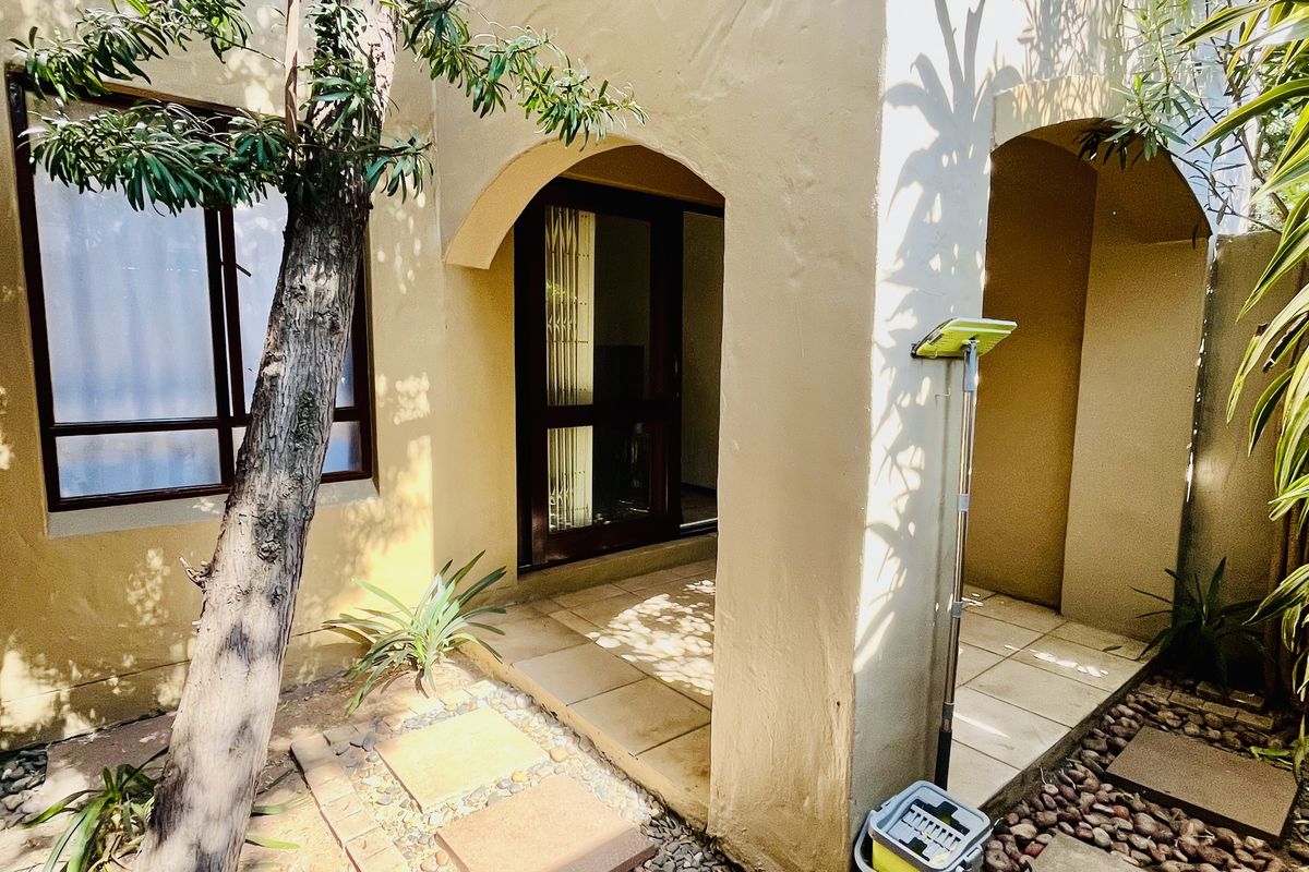 A rear exterior view of the apartment featuring a covered tiled patio, arched architectural details, and a landscaped garden area with stone pathways.