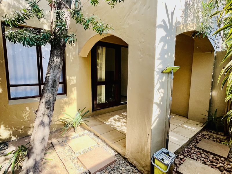 A rear exterior view of the apartment featuring a covered tiled patio, arched architectural details, and a landscaped garden area with stone pathways.