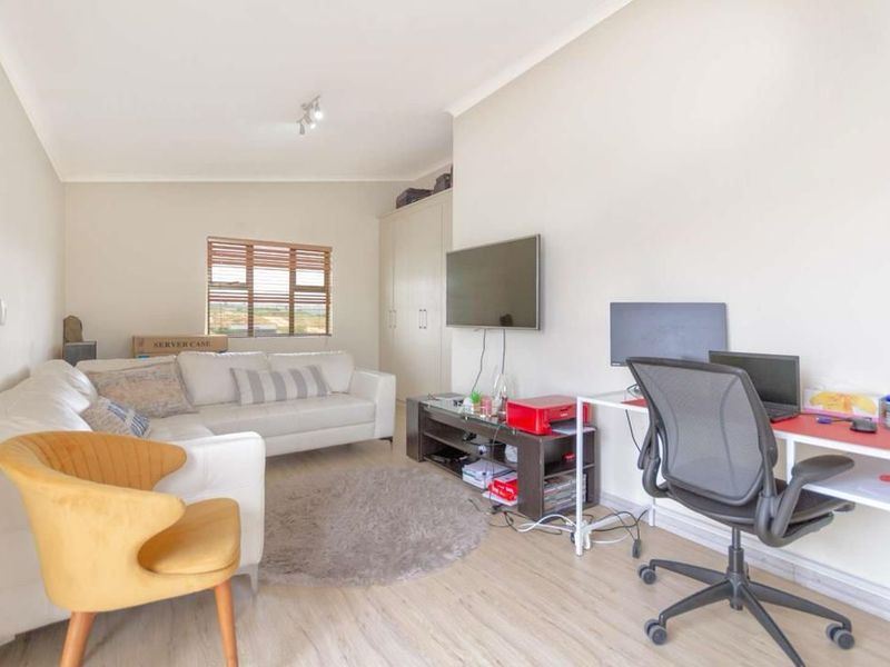 A functional living room featuring laminate flooring, built-in storage cupboards, and window blinds, currently utilized as a combined lounge and home office space.