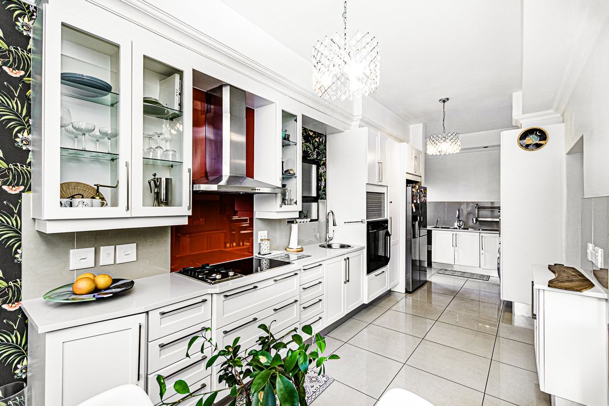 Modern kitchen featuring white cabinetry, stone countertops, a striking red glass splashback, built-in appliances, and tiled flooring, with an adjacent scullery area.