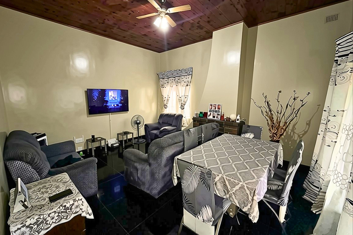 A living and dining area featuring a dark timber-clad ceiling, ceiling fan, and glossy floor tiles, providing a functional space for relaxation and meals.