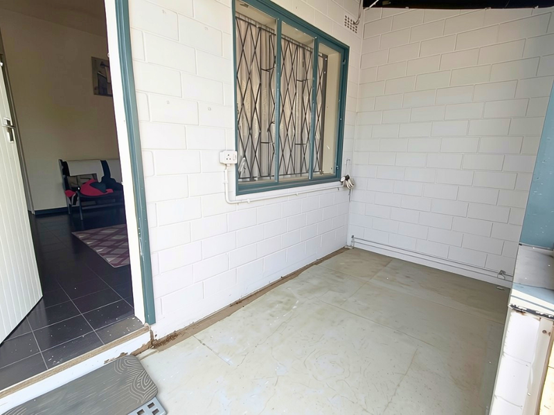 A covered patio area featuring white painted brick walls, security window bars, and a concrete floor, providing a functional outdoor transition space.