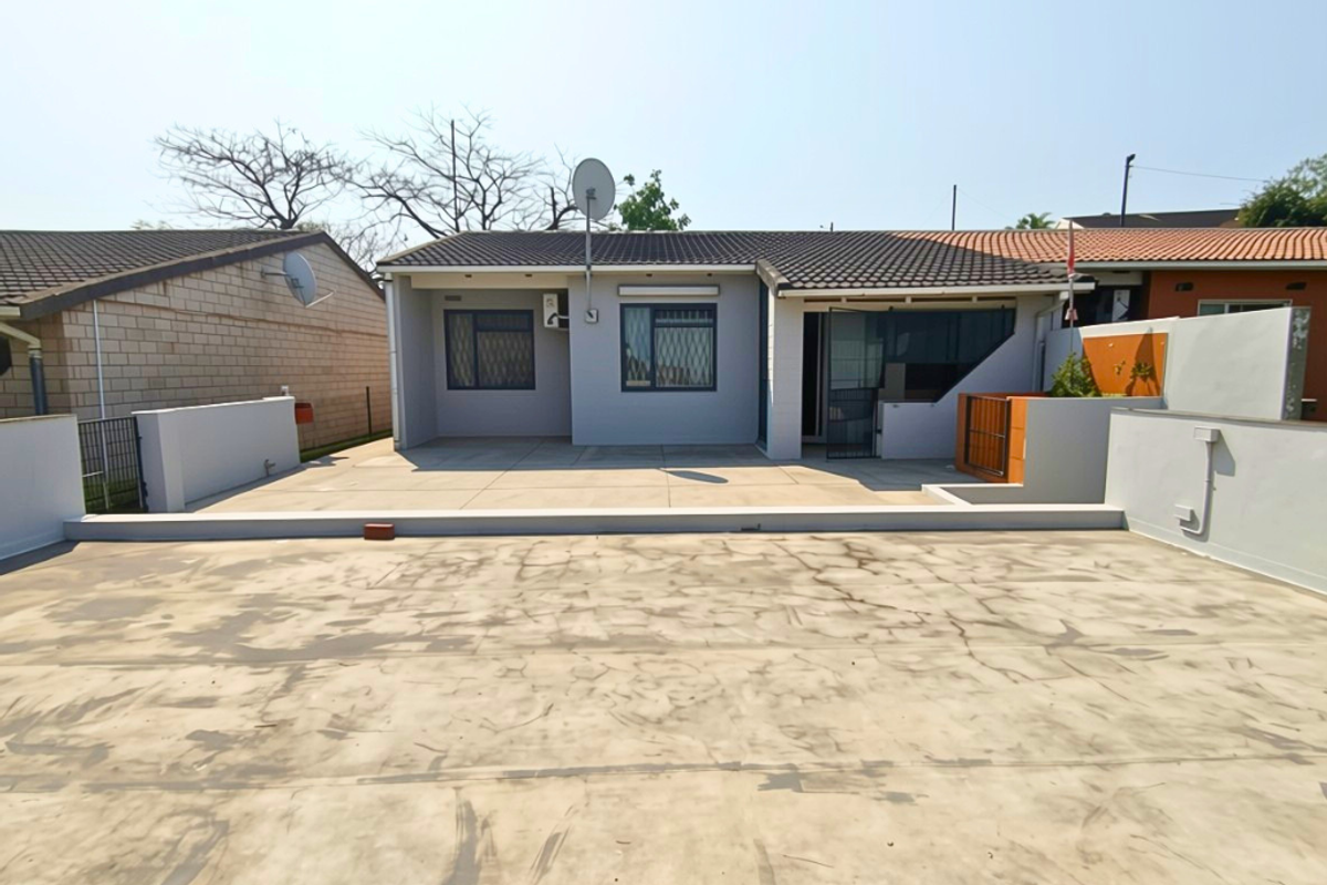 A rear exterior view of the house featuring a large concrete patio area, tiled roof, and perimeter boundary walls.