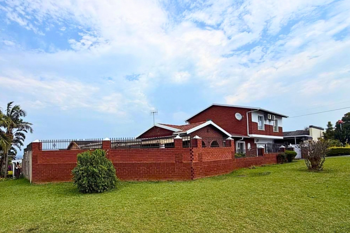 A view of the rear exterior of the house featuring a large lawn, brick boundary wall with metal fencing, and a tiled roof structure.