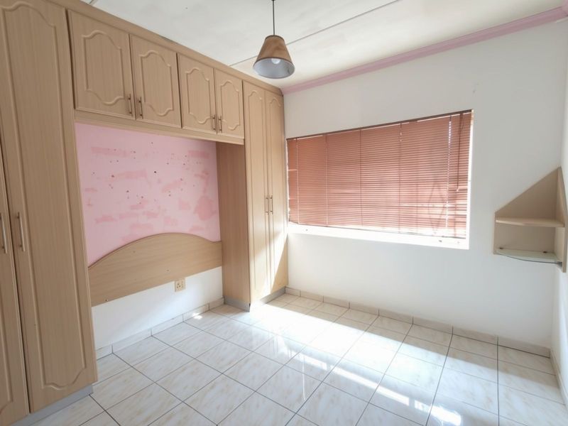 A vacant bedroom featuring built-in wooden cupboards, tiled flooring, and a window with venetian blinds. The room requires minor cosmetic repairs to the wall.