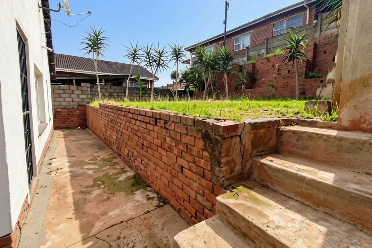 Rear exterior area featuring a brick retaining wall, concrete stairs, and paved ground, showing signs of wear and structural cracking.