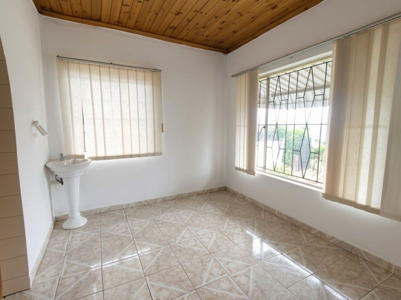 A bright, empty bedroom featuring tiled flooring, a timber ceiling, and a built-in pedestal washbasin with security bars on the windows.
