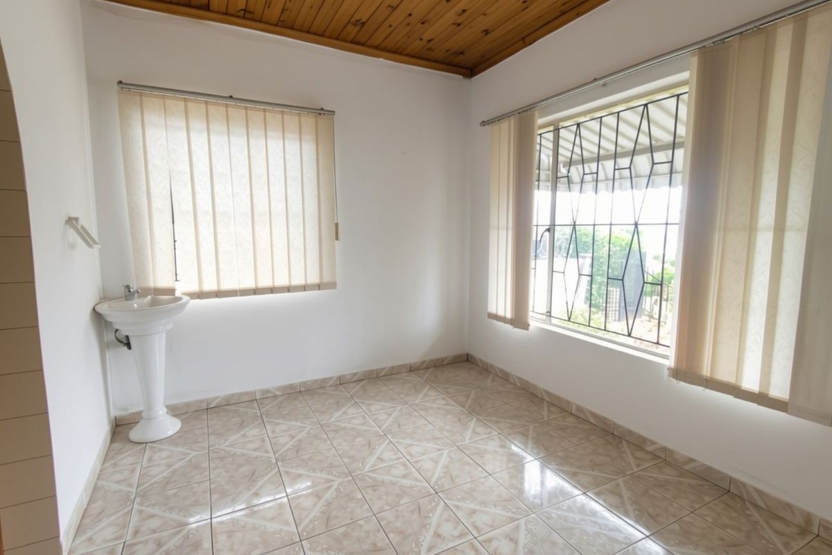 A bright, empty bedroom featuring tiled flooring, a timber ceiling, and a built-in pedestal washbasin with security bars on the windows.