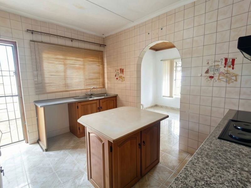 A functional kitchen featuring wooden cabinetry, a central island, double sink, and tiled finishes, offering a classic layout with an adjoining room through an archway.
