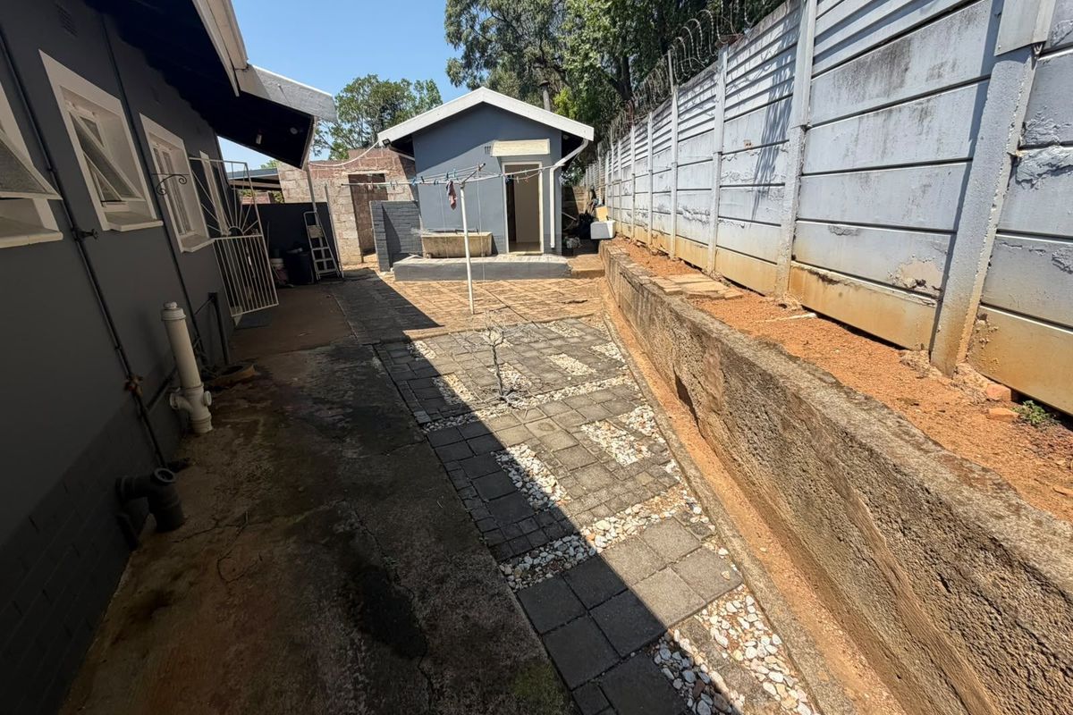 Rear exterior view showing a paved walkway, retaining wall, and a detached outbuilding with a precast concrete boundary wall and security fencing.