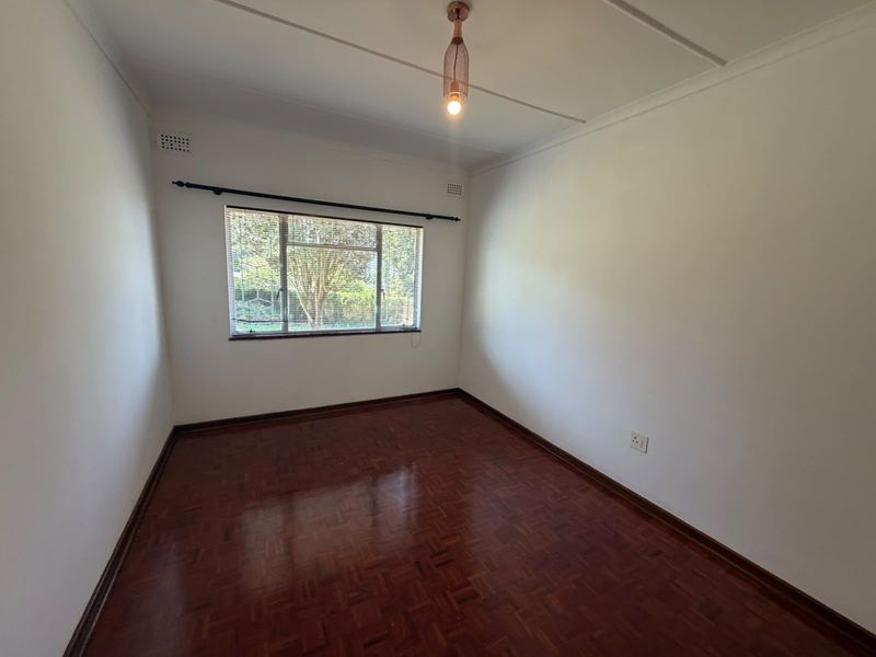 A bright, empty bedroom featuring polished parquet flooring, a single pendant light fixture, and a window with fitted blinds.