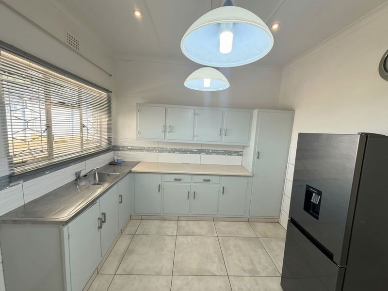 A functional kitchen featuring light-toned built-in cabinetry, a stainless steel sink, tiled backsplash, and neutral floor tiles with ample natural light.