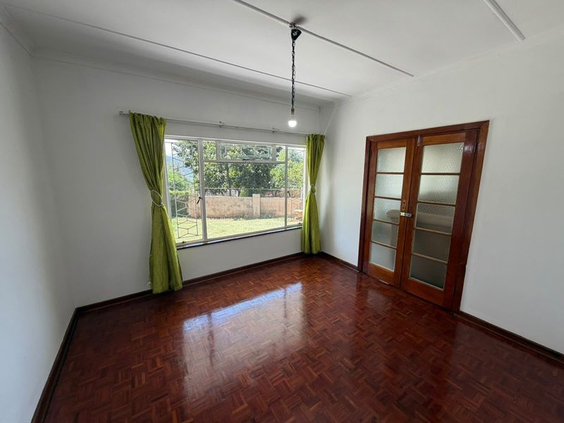 A bright, empty living room featuring polished parquet flooring, wooden double doors with glass panels, and a large window overlooking the garden.