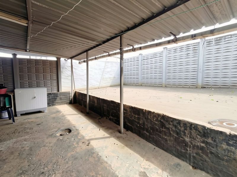 A covered outdoor patio area featuring a corrugated metal roof, brick retaining wall, and concrete flooring, enclosed by precast concrete boundary walls.