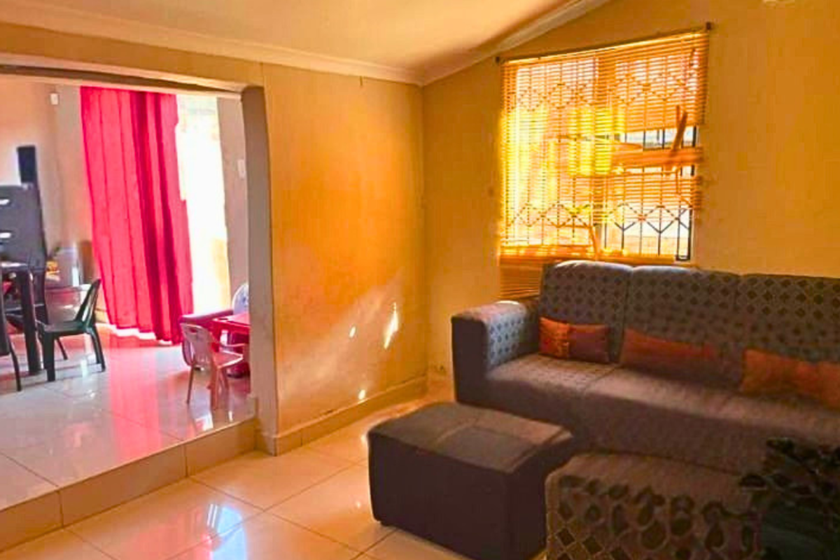A living room featuring tiled flooring and an open-plan layout connecting to an adjacent space, with natural light filtering through window blinds.