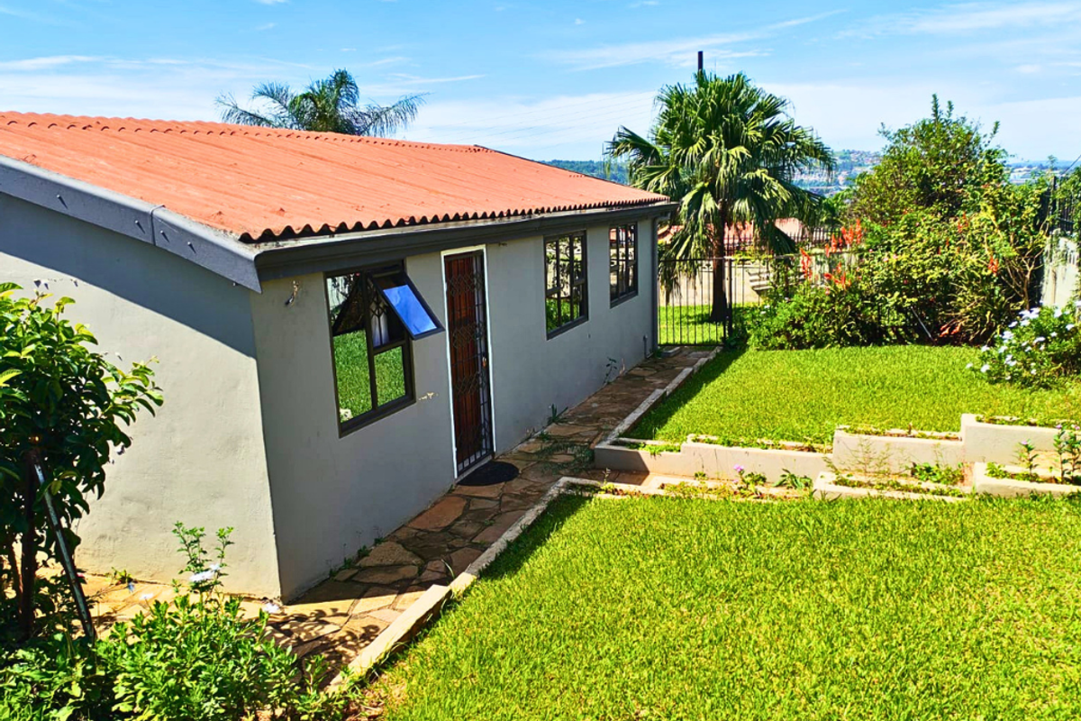 A view of the rear exterior of the house featuring a corrugated roof, paved walkway, and a well-maintained lawn with landscaped garden beds.