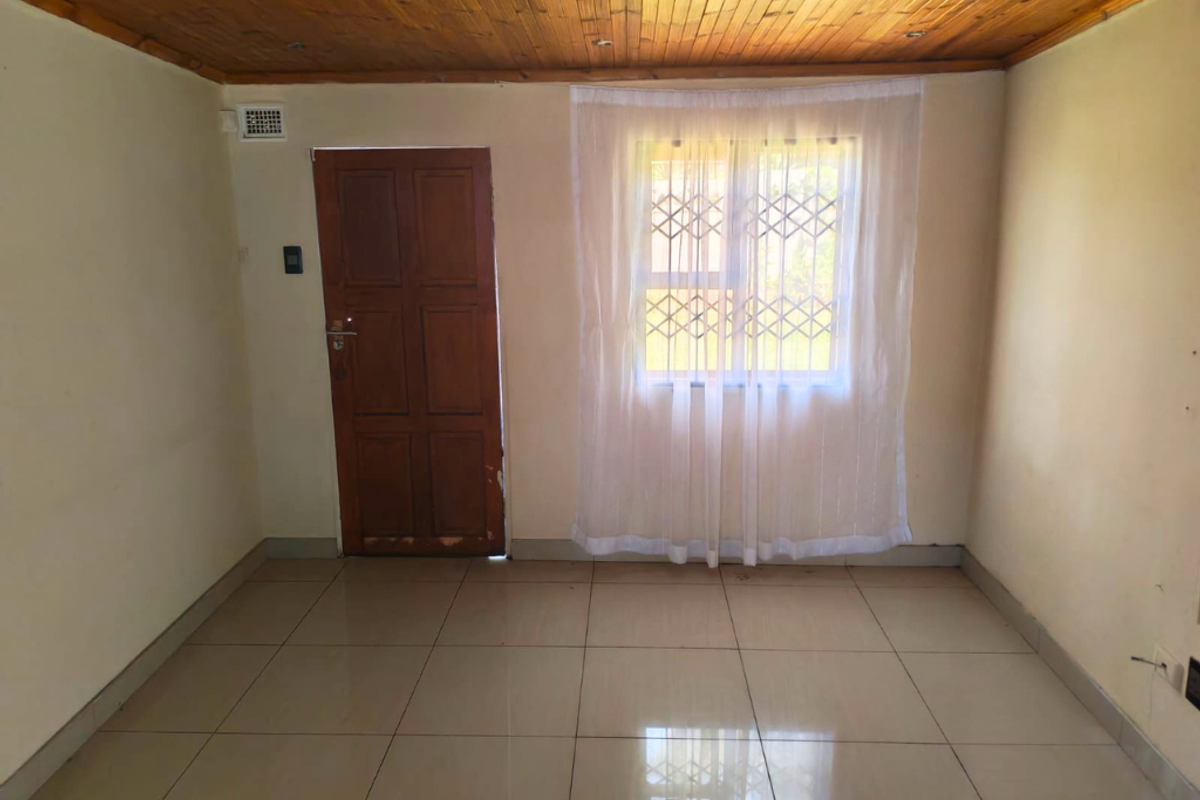 A simple, tiled living room featuring a wooden ceiling, a solid timber door, and a window fitted with security bars.