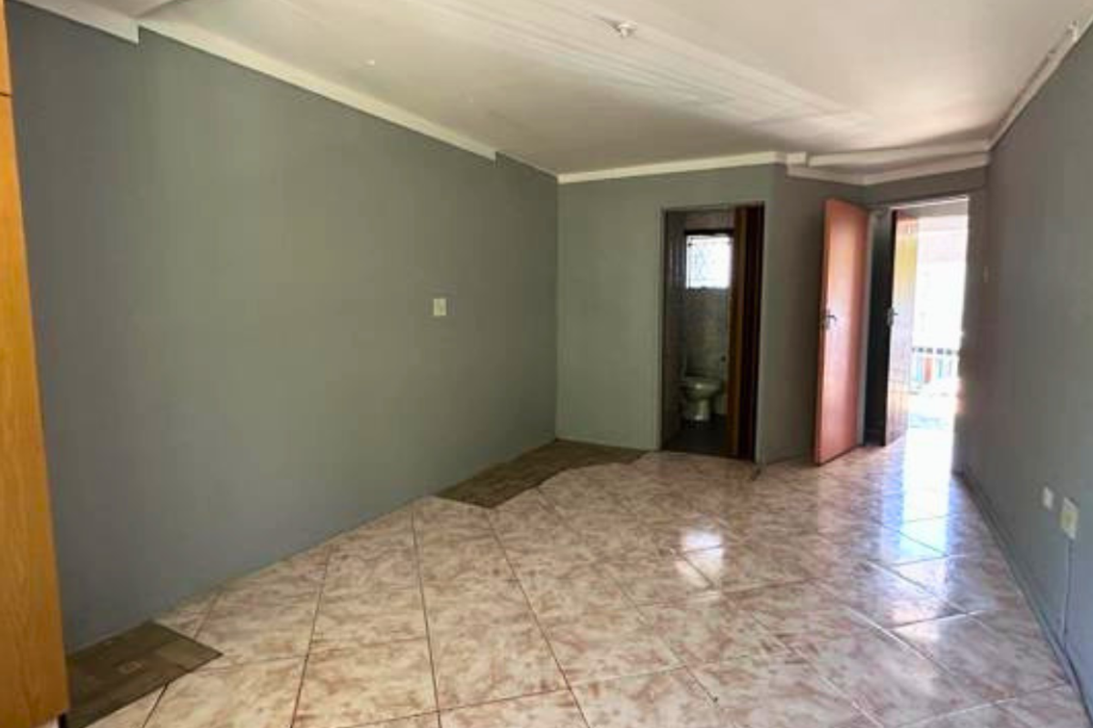 An empty living area featuring tiled flooring, neutral grey walls, and direct access to an adjoining bathroom and exterior doorway.