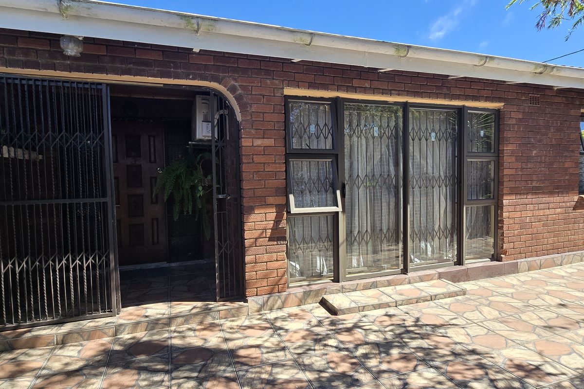 A rear exterior view of a brick-faced house featuring a paved patio area, security gates, and aluminium-framed windows.