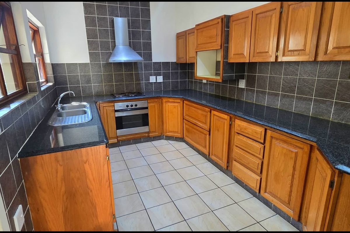 A functional kitchen featuring wooden cabinetry, granite countertops, a built-in oven and hob, and a stainless steel extractor fan with tiled finishes.