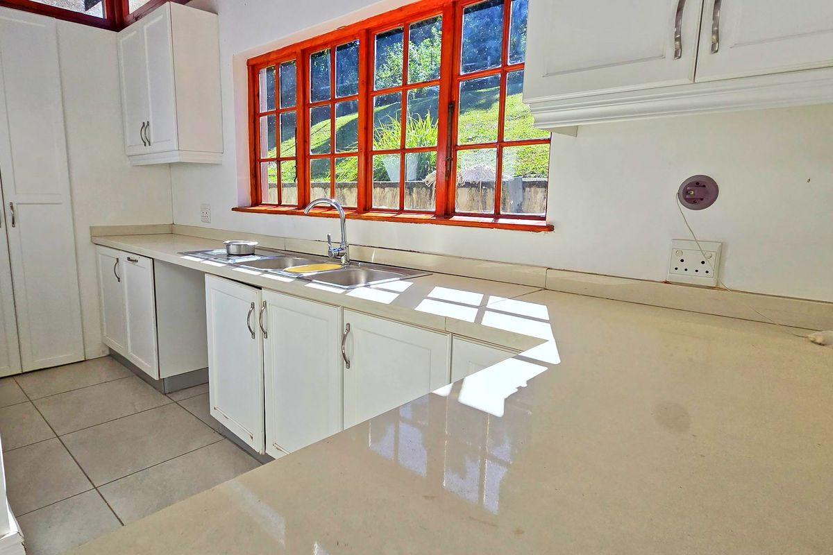 A bright, empty kitchen featuring white cabinetry, stone countertops, a stainless steel sink, and large timber-framed windows overlooking the garden.