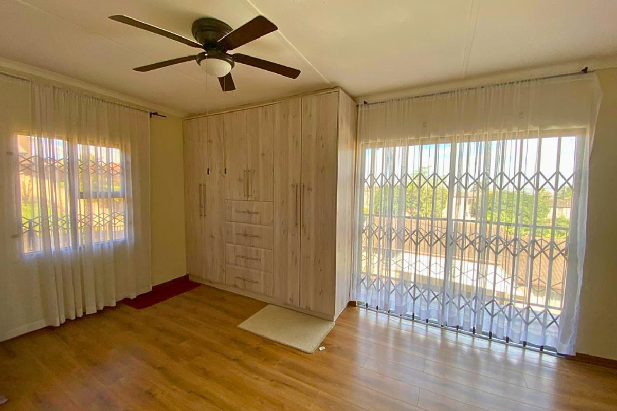 Spacious bedroom featuring light wood-finish built-in wardrobes, laminate flooring, a ceiling fan, and sliding glass doors with security gates leading to the exterior.