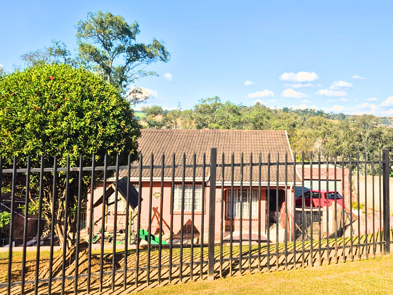 A single-story residential house featuring a tiled roof, brick facade, and a secure metal perimeter fence with a driveway and attached garage.
