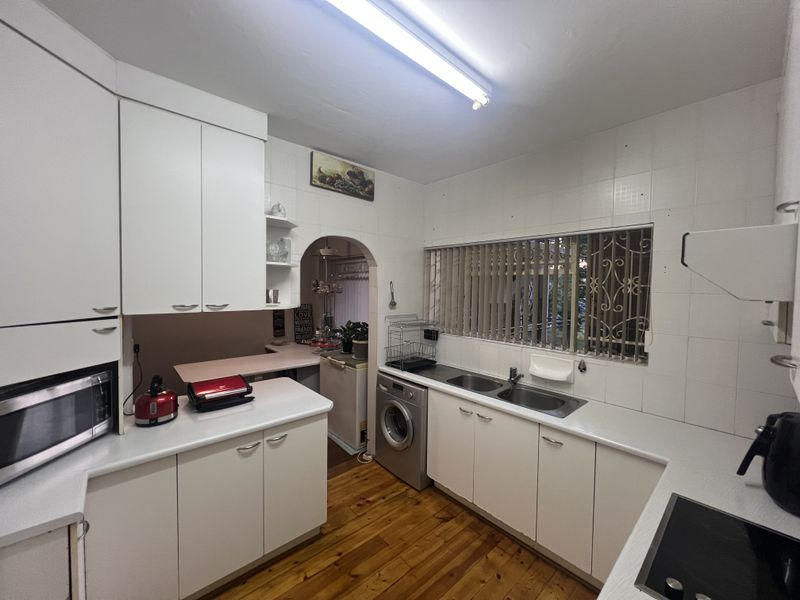 A functional kitchen featuring white cabinetry, laminate countertops, a double basin sink, and integrated laundry plumbing, with an arched doorway leading to a scullery area.