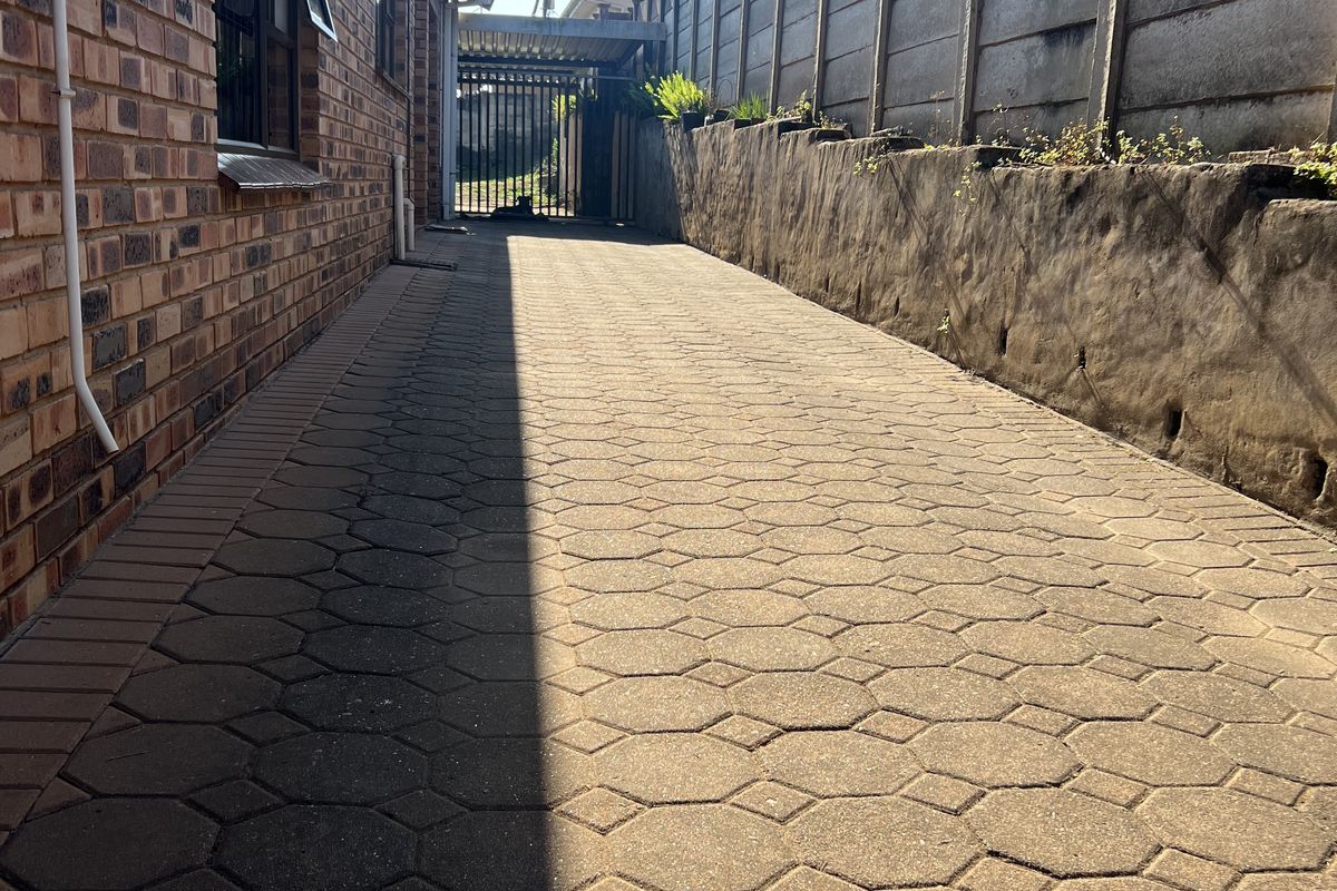 A paved side walkway featuring a face-brick exterior wall and a concrete boundary wall, leading towards a gated entrance.