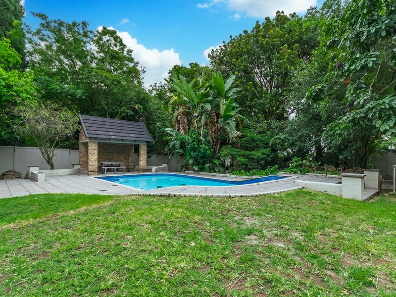 Outdoor pool area featuring a swimming pool, paved surround, and a brick gazebo set within a landscaped garden with mature trees.