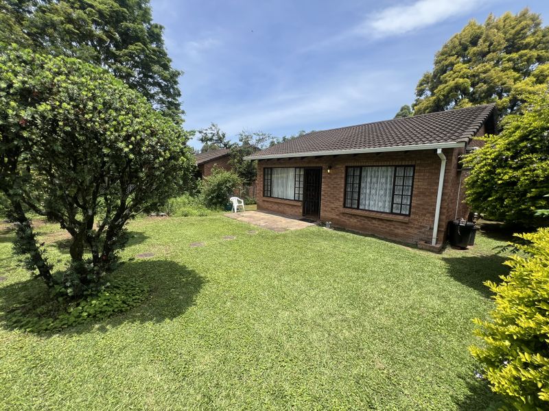 A rear view of a brick townhouse featuring a small paved patio area and a spacious, established garden with mature trees.