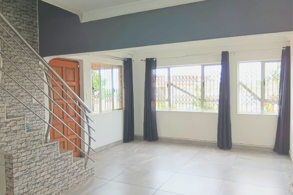 Spacious living area featuring tiled flooring, a decorative stone-clad staircase with metal railings, and large windows with security bars.