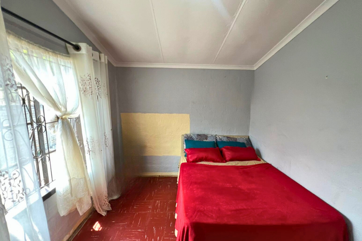 A bedroom featuring red tiled flooring, grey painted walls with a patch repair, and a window fitted with security bars.