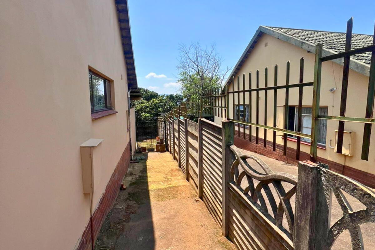 A narrow side passage between two residential structures featuring concrete boundary fencing, security spikes, and paved ground surface.