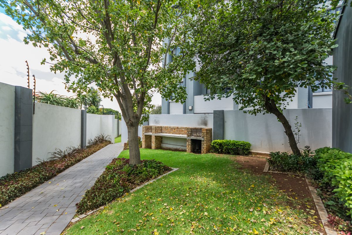 A private rear garden featuring a paved walkway, established trees, and a brick built-in braai area enclosed by a perimeter wall with electric fencing.