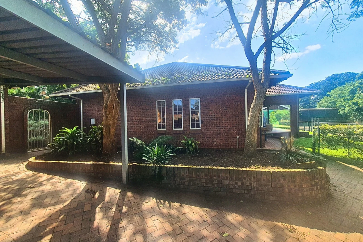 Exterior view of a brick house featuring a covered patio area, paved driveway, and established garden beds with mature trees.