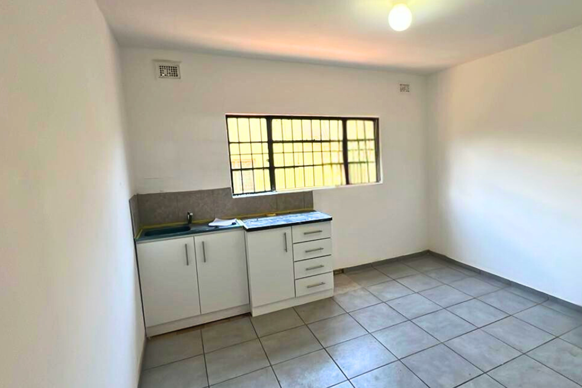 A compact kitchen area featuring white cabinetry, tiled flooring, and a window with security bars, offering a functional space ready for customization.