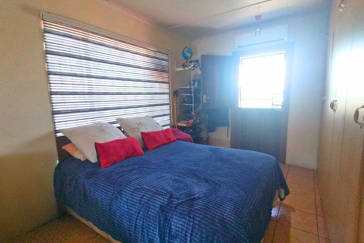 A bedroom featuring tiled flooring, built-in cupboards, a wall-mounted air conditioning unit, and a large window with fitted blinds.