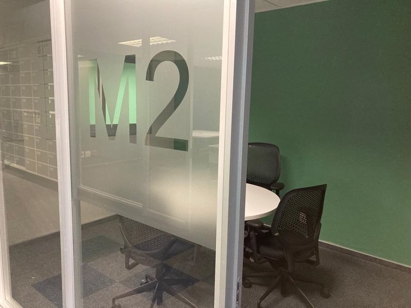 A small private office meeting room featuring a frosted glass partition, green accent wall, and carpeted flooring.