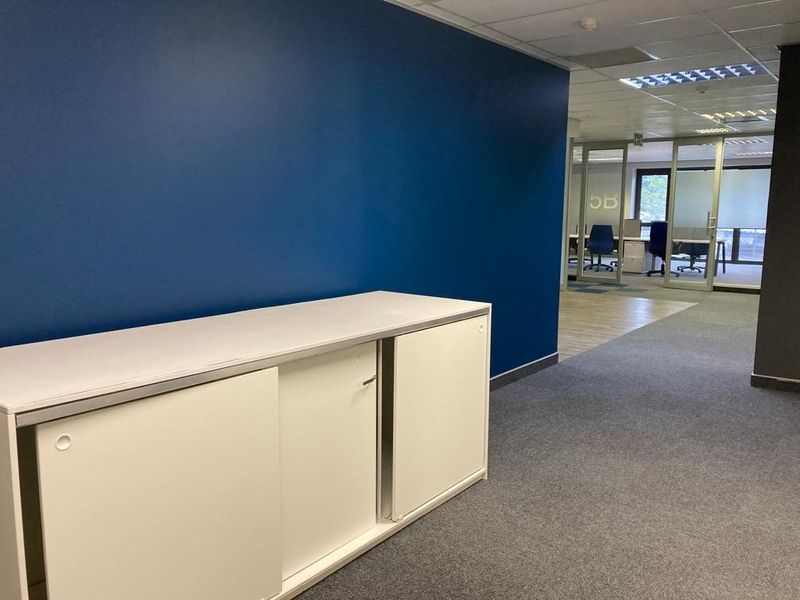 Modern office space featuring glass partitions, carpeted flooring, recessed lighting, and a built-in storage unit against a blue accent wall.