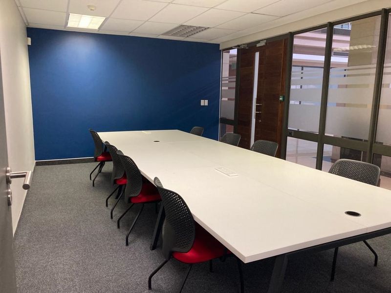 A modern office meeting room featuring a large central table, carpeted flooring, a blue accent wall, and glass partition walls for a professional workspace.